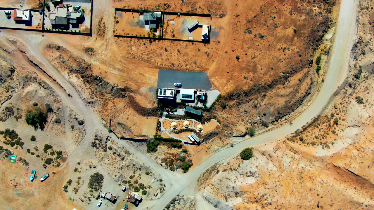 High aerial frame over San Juanico showing graded dirt roads, fenced or walled home parcels, a paved driveway, orange soil, scrub patches, and no visible people.
