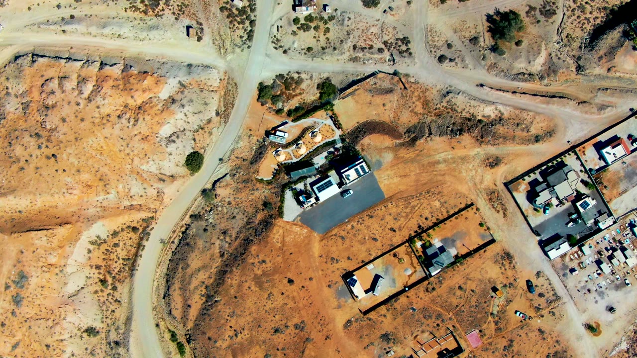 Near-vertical aerial view over San Juanico, Baja California Sur, showing sandy desert terrain, dirt roads, walled lots, modern buildings, parked vehicles, and no visible people.