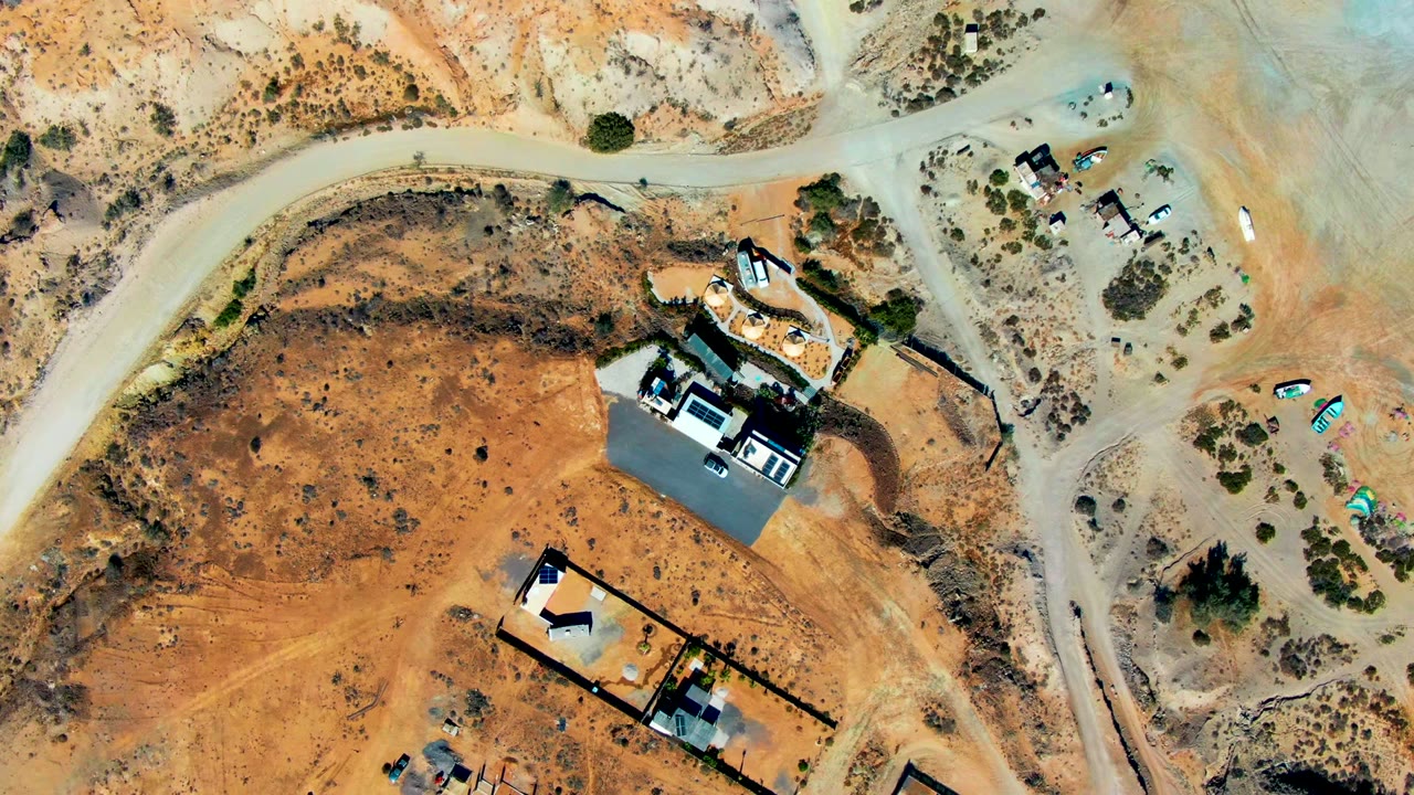 Top-down San Juanico aerial frame showing a broad sandy building area, angular walled properties, dirt access roads, scattered vehicles, small shrubs, and no visible people.