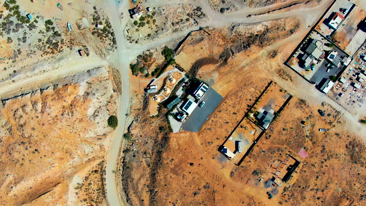 Overhead aerial frame in San Juanico, Baja California Sur showing intersecting dirt roads, walled construction parcels, sparse scrub, parked vehicles, and no visible people.