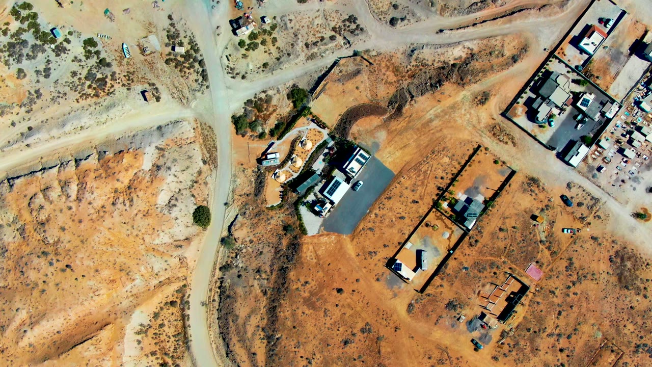 Aerial frame over San Juanico, Baja California Sur showing pale dirt roads crossing dry terrain, several developed parcels, small structures, vehicle clusters, and no visible people.