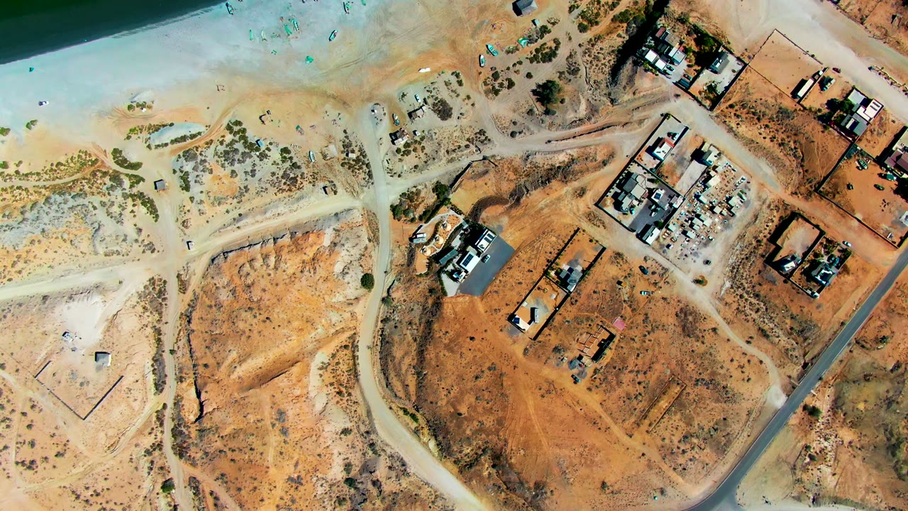 Top-down San Juanico, Baja California Sur frame showing intersecting dirt roads, orange-brown construction lots, a cluster of buildings at the upper right, a paved road along the right side, and no visible people.