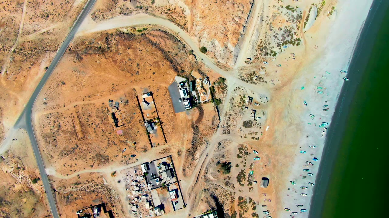Overhead aerial frame in San Juanico, Baja California Sur showing dry orange lots, dirt tracks, a paved road, buildings, pale beach sand, green shoreline water, and no visible people.