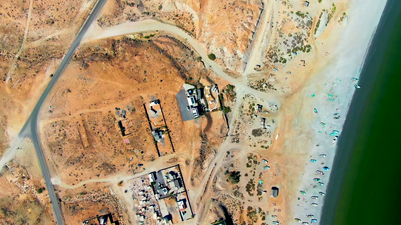 Aerial view of a coastal road, buildings, sandy beach, and small umbrellas.