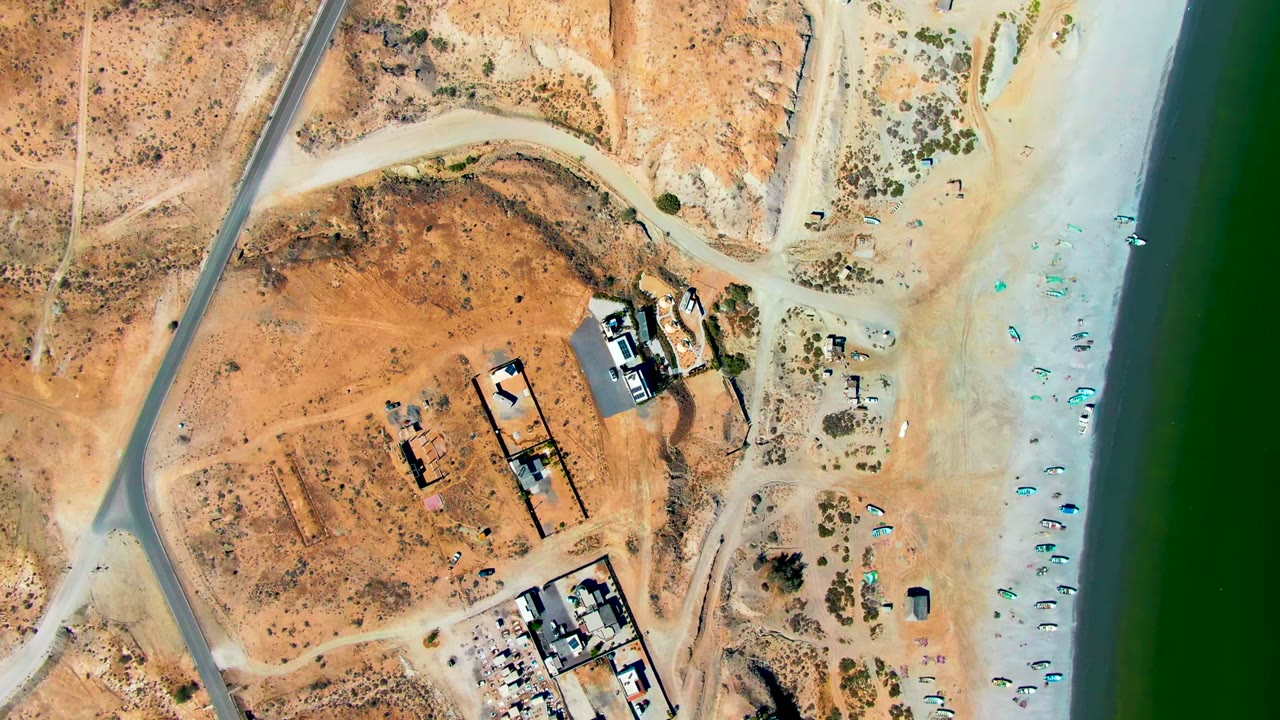 Top-down aerial frame in San Juanico, Baja California Sur showing a block of buildings, empty dry ground, pale beach, rows of small umbrellas near green water, and no visible people.