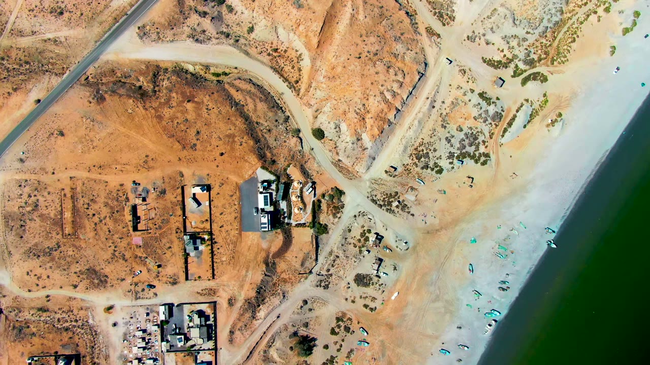 Overhead view in San Juanico, Baja California Sur showing sandy lots, low buildings, beach gear, tiny visible people near the shore, and dark green water along the right edge.