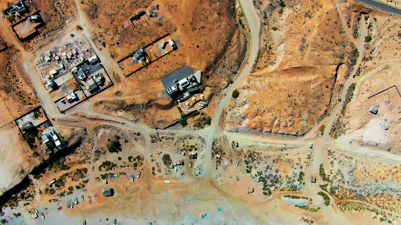 Aerial overhead in San Juanico with pale dirt roads crossing dry lots, a few low buildings, scrub patches, parked vehicles, and small beach details near the bottom edge.