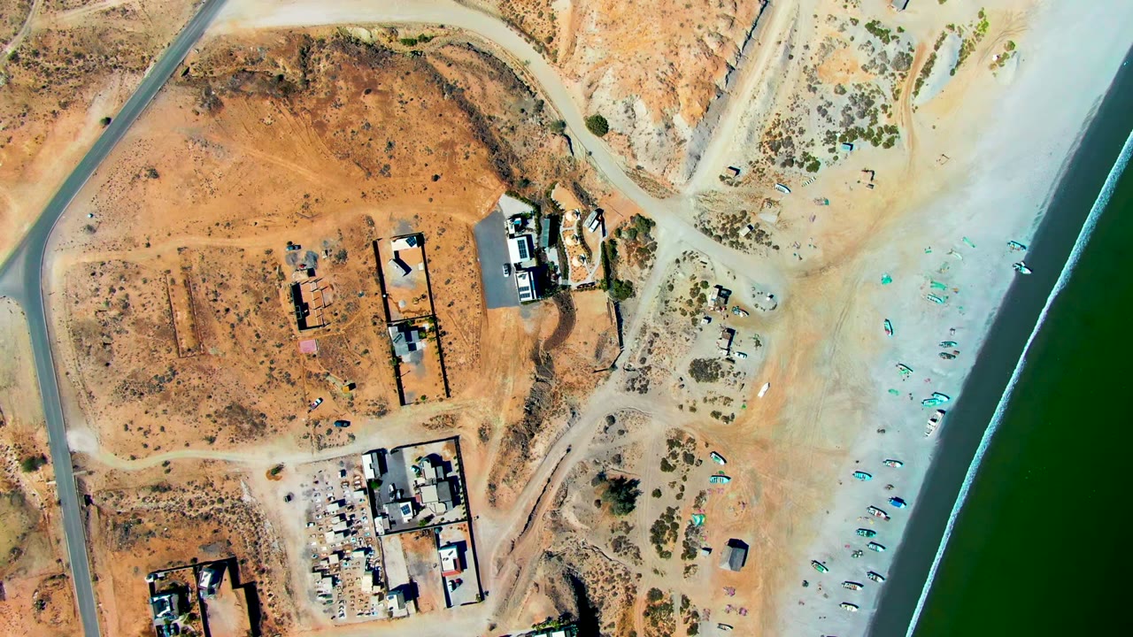 Aerial frame over San Juanico, Baja California Sur, showing tan dirt lots, unfinished buildings, pale beach flats, green water along the right edge, and no visible people.