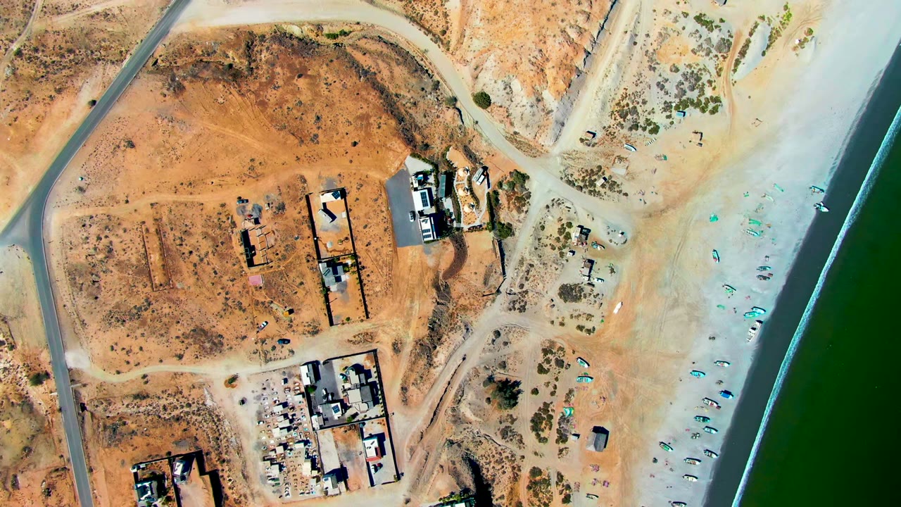 Top-down San Juanico aerial with the road curving at left, construction buildings near center, pale shoreline sand to the right, green water beyond it, and no visible people.