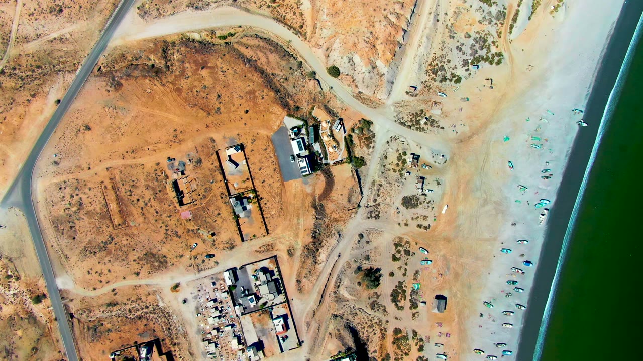 San Juanico aerial frame showing dusty roads and graded lots on the left, clustered construction structures near the middle, small blue shore objects, green water at right, and no visible people.