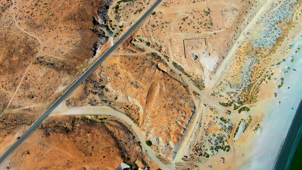 Top-down aerial frame in San Juanico, Baja California Sur showing a paved road, tan desert terrain, dirt access tracks, pale beach, green water, and tiny beach users near the shoreline.