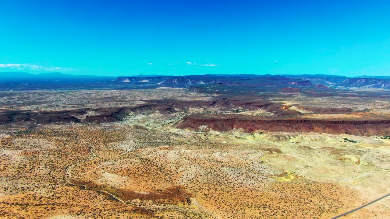 Wide aerial view over San Juanico desert terrain in Baja California Sur, with sparse scrub, pale washes, low distant mountains, clear blue sky, and no visible people.