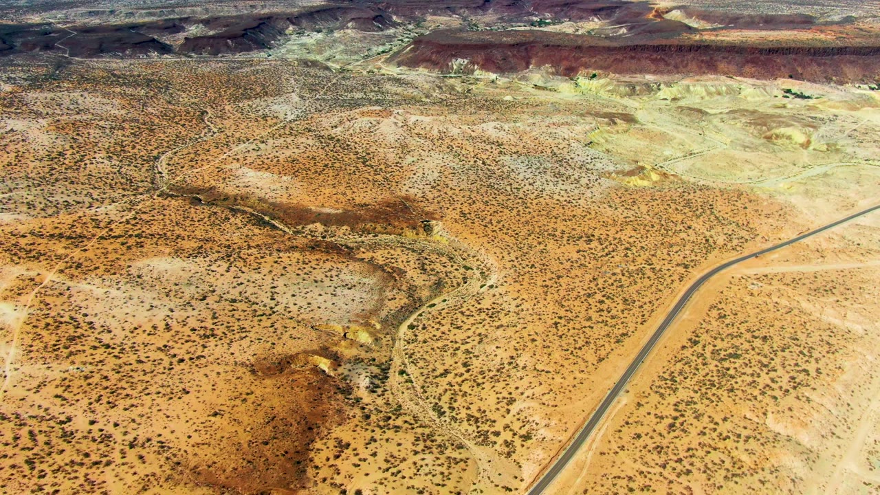 Steeper drone view over San Juanico desert scrub and a winding dry wash, with a straight paved road crossing the right side, pale hills beyond, and no visible people.