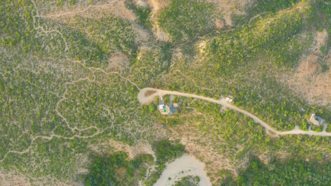 Overhead aerial frame in Cabo Pulmo, Baja California Sur, showing scrub vegetation, a curved dirt road, small structures near the road, and no visible people.