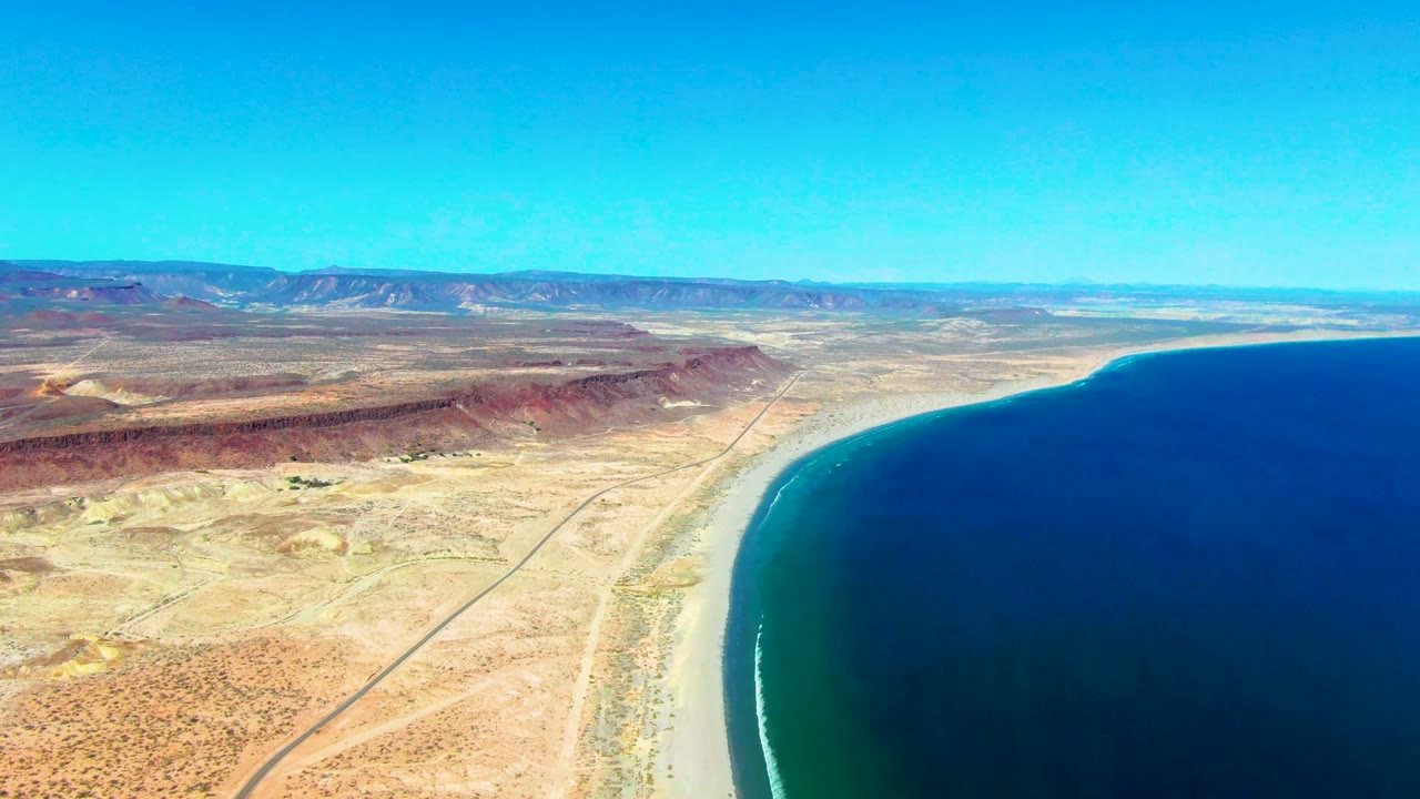 Aerial view over San Juanico, Baja California Sur with deep blue ocean at right, a pale sandy beach below, desert road at left, distant mountains, and no visible people.