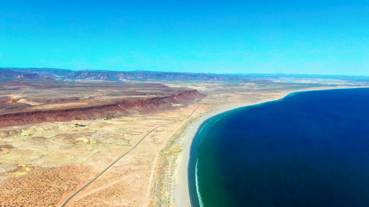 Aerial coastline with ocean, beach curve, dry desert plain, road, and mountain horizon.