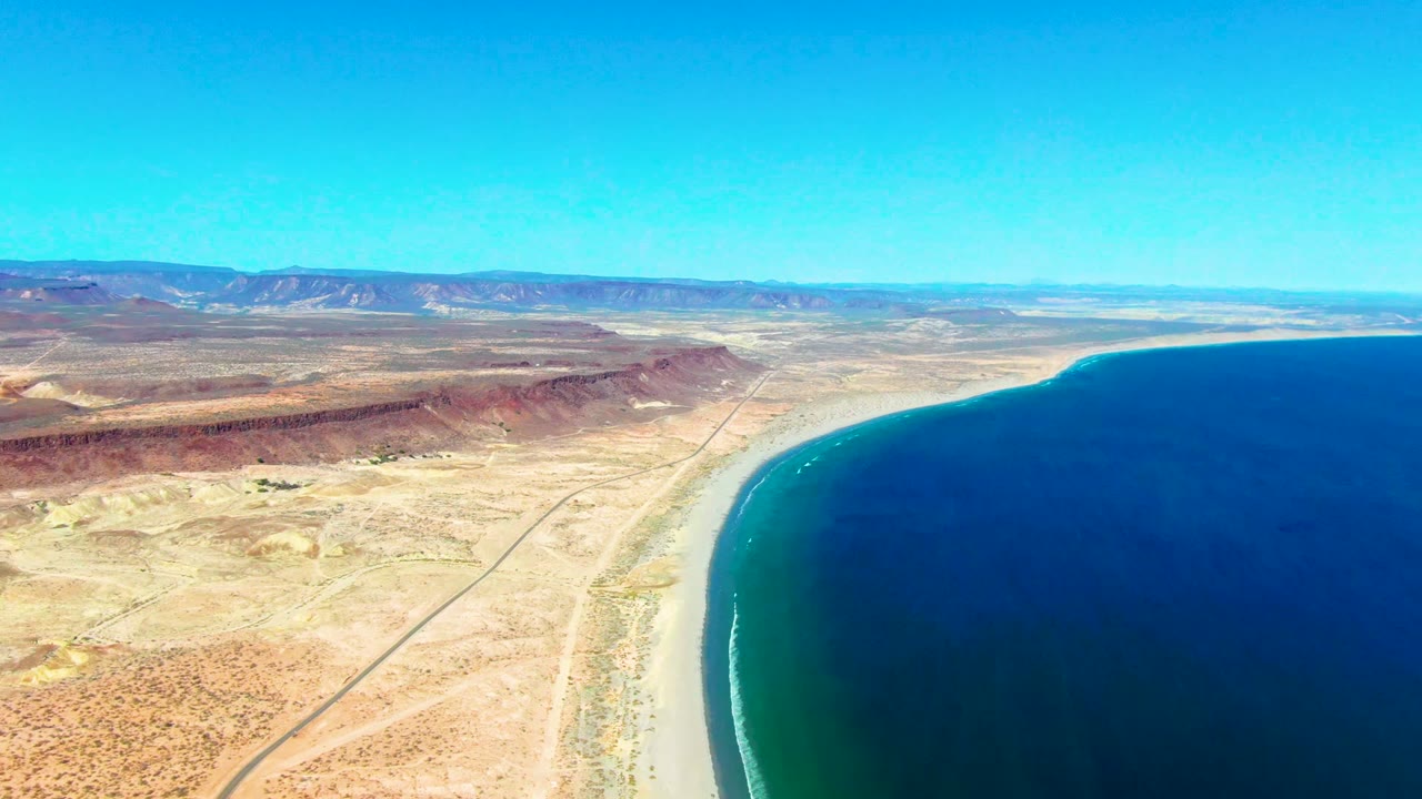 San Juanico coastal aerial with the ocean filling the right side, a narrow beach strip below, red-brown inland hills, a road crossing the desert, and no visible people.