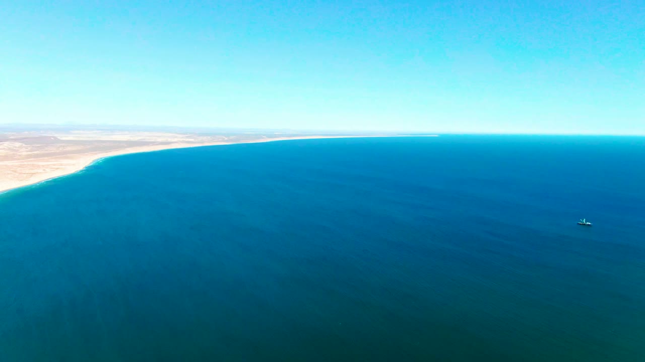 Wide aerial view over San Juanico, Baja California Sur, with blue ocean filling most of the frame, desert coast at left, clear sky, and no visible people.