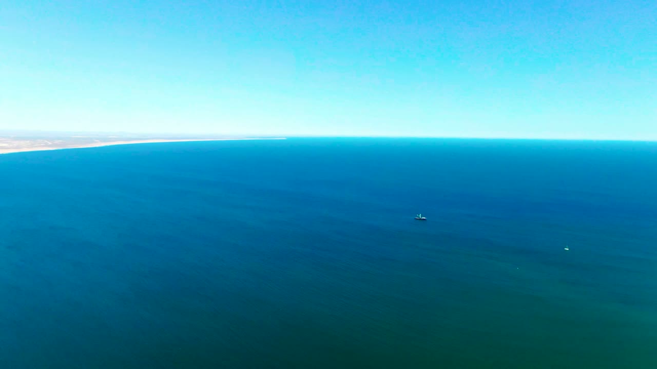 Aerial view near San Juanico, Baja California Sur, showing open blue water, a small pale sandbar or exposed coastal feature, distant vessels, clear sky, and no visible people.