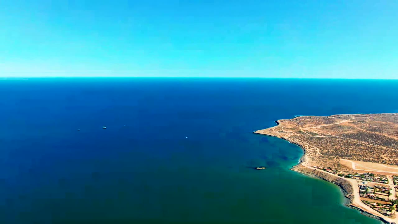 Aerial view over San Juanico, Baja California Sur, with dry coastline on the right, blue ocean filling the left side, clear sky, and no visible people.