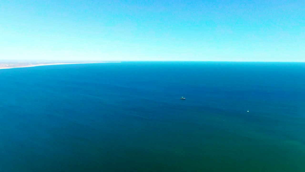 Aerial view of deep blue ocean near San Juanico, with a straight horizon, faint coastline at the far left, small distant surface specks, and no visible people.