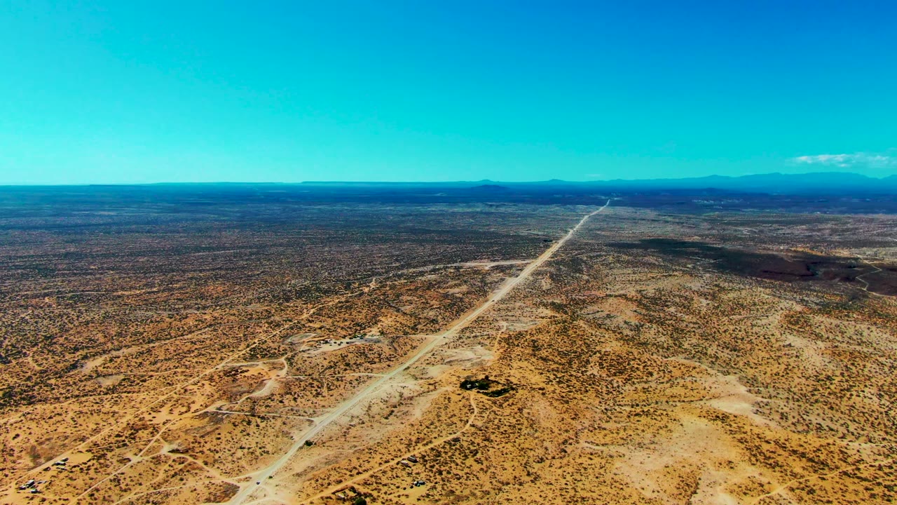 High aerial view over San Juanico, Baja California Sur, with a long dirt road cutting through tan desert scrub toward a distant blue coastline and no visible people.