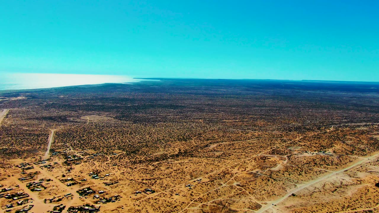Wide aerial view of San Juanico with scrubland, pale dirt roads, scattered low buildings, and the coastal waterline across the background, with no visible people.