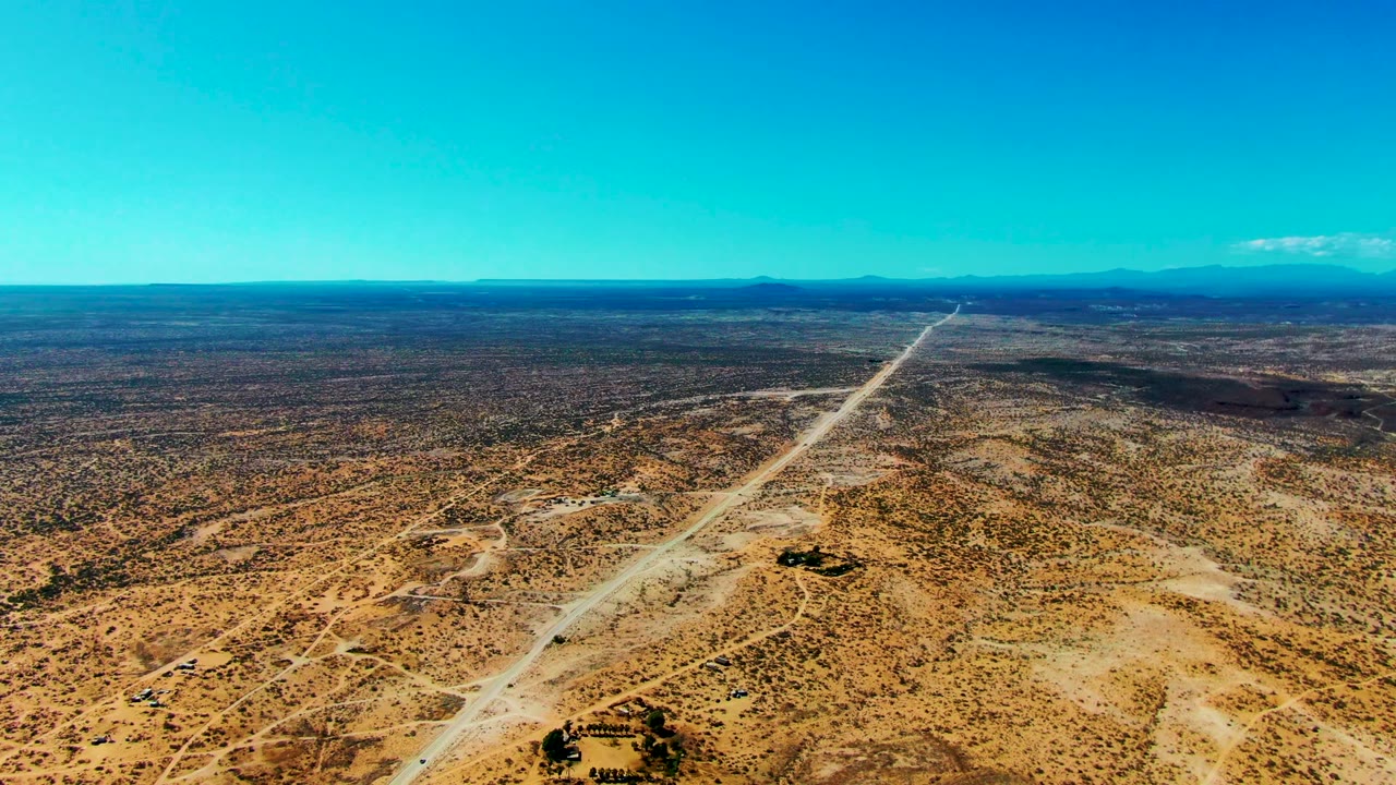 High aerial frame of San Juanico desert terrain where the pale road runs from the lower frame toward the mountain horizon, with sparse brush and no visible people.