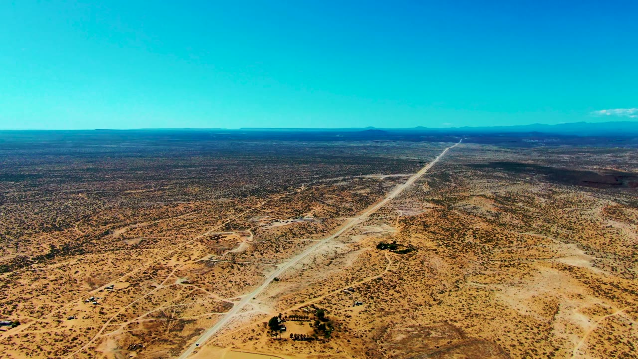 Wide San Juanico aerial with the pale road running diagonally through beige and brown desert, distant mountains on the horizon, and no visible people.