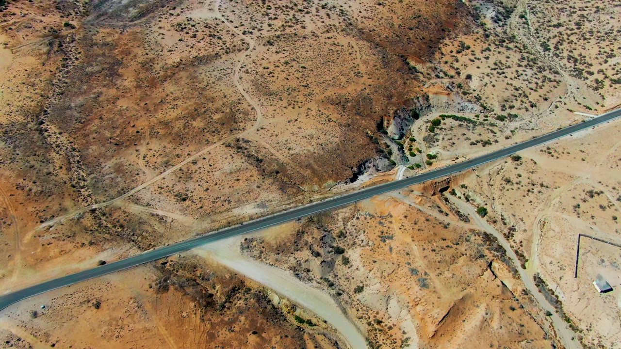 Aerial view over San Juanico desert terrain with a paved road cutting across tan hills, dirt pullouts, scrub, small structures, and no visible people.