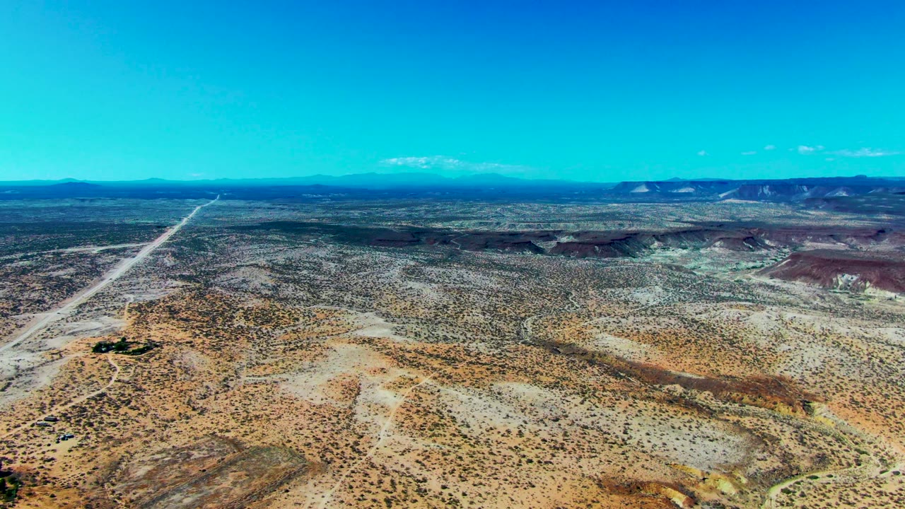 High aerial panorama over San Juanico desert with a straight dirt road receding toward the horizon, low scrub, layered ridges, clear blue sky, and no visible people.