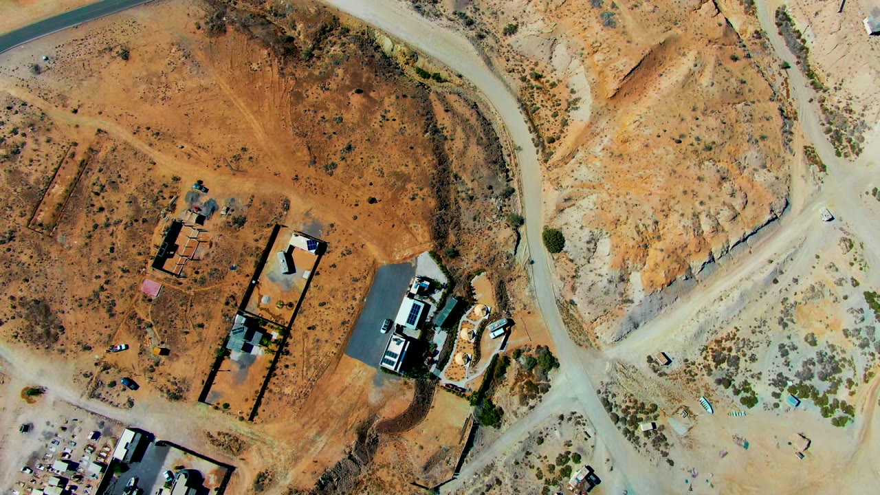 Overhead drone frame of a San Juanico desert construction site with reddish earth, pale dirt roads, unfinished structures, parked vehicles, scrub, and no visible people.