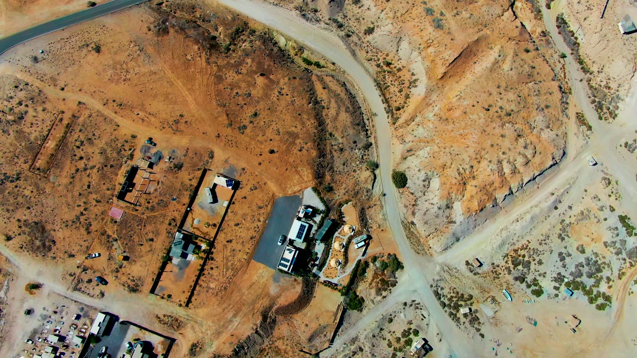 Aerial frame in San Juanico with a paved road near the top, intersecting dirt tracks, reddish construction ground, scattered buildings, and no visible people.