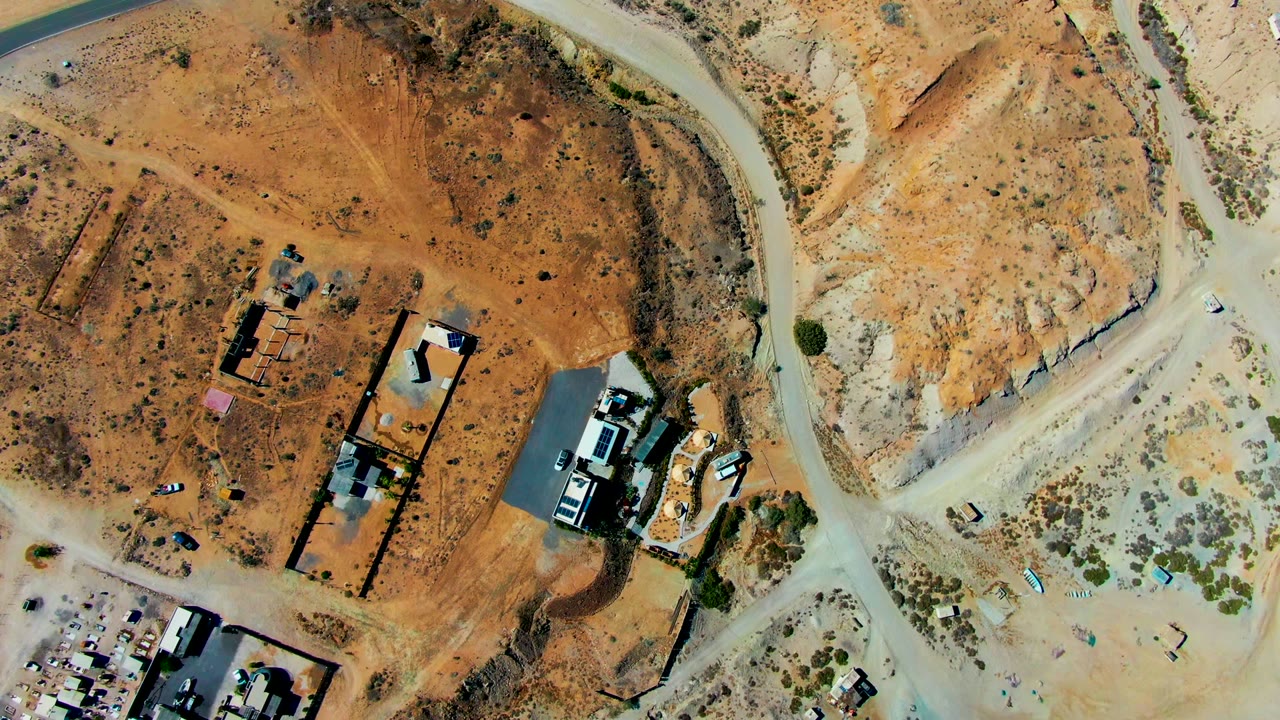 Overhead drone frame of a San Juanico construction area with orange dirt ground, pale roads, sparse shrubs, several buildings, and no visible people.