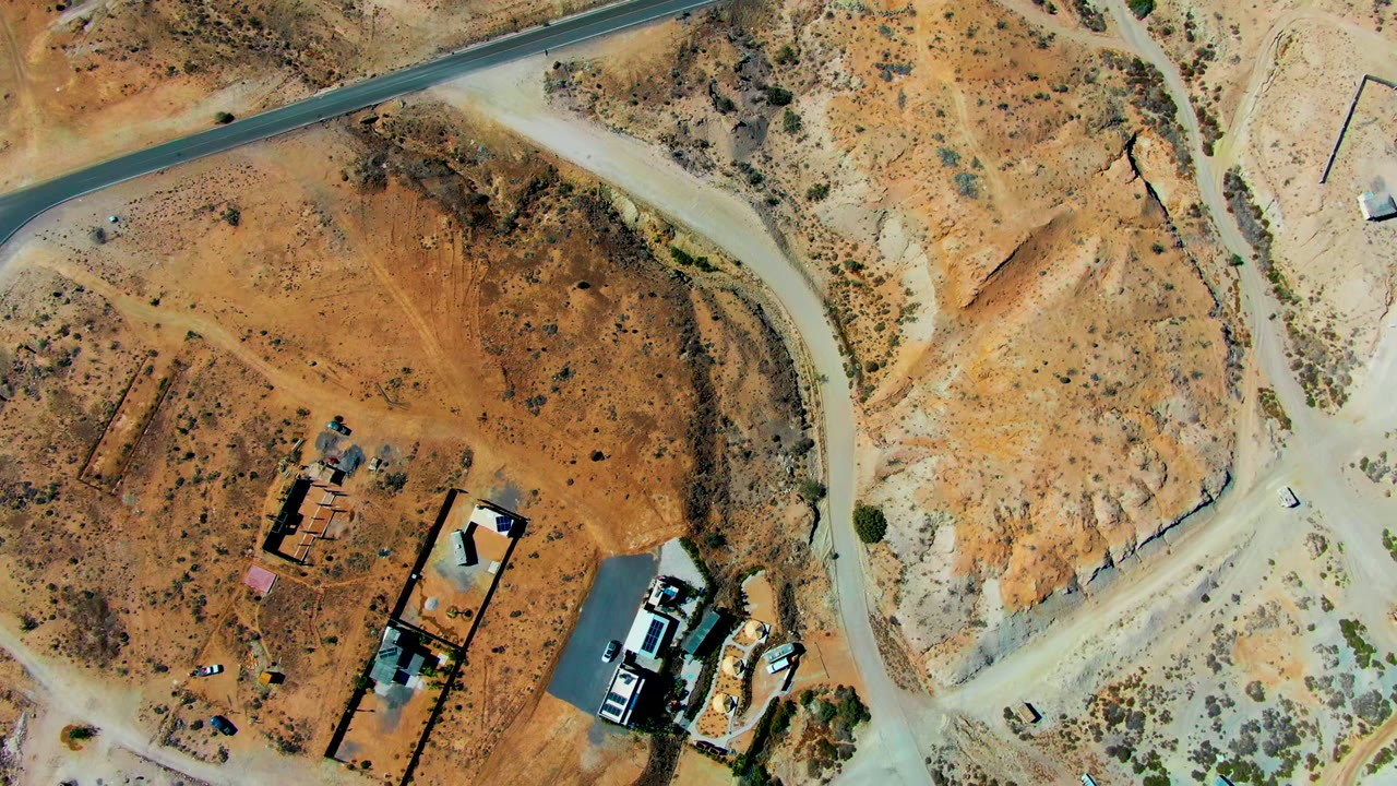 Top-down aerial frame over San Juanico, Baja California Sur showing dry orange terrain, intersecting pale roads, scattered small structures, vehicles, and no visible people.