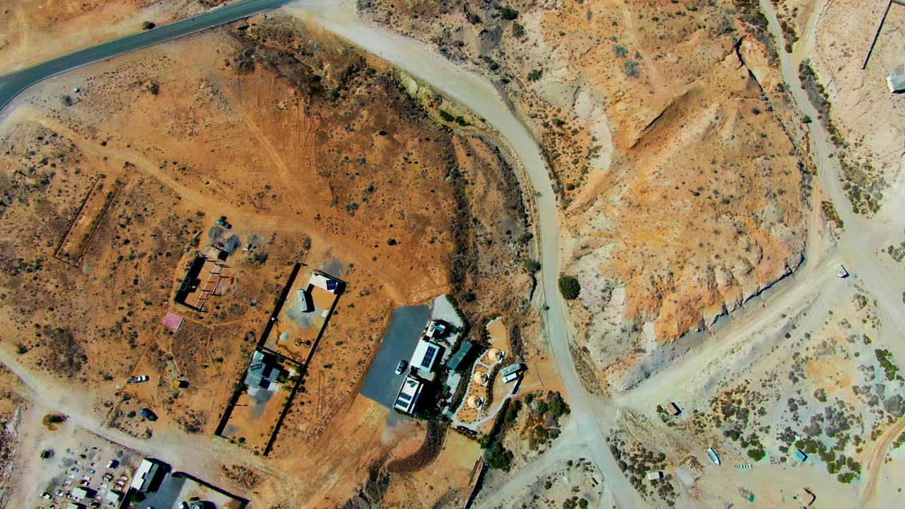 Aerial frame in San Juanico with a pale road junction beside orange desert lots, several small buildings, light-colored vehicles, and no visible people.