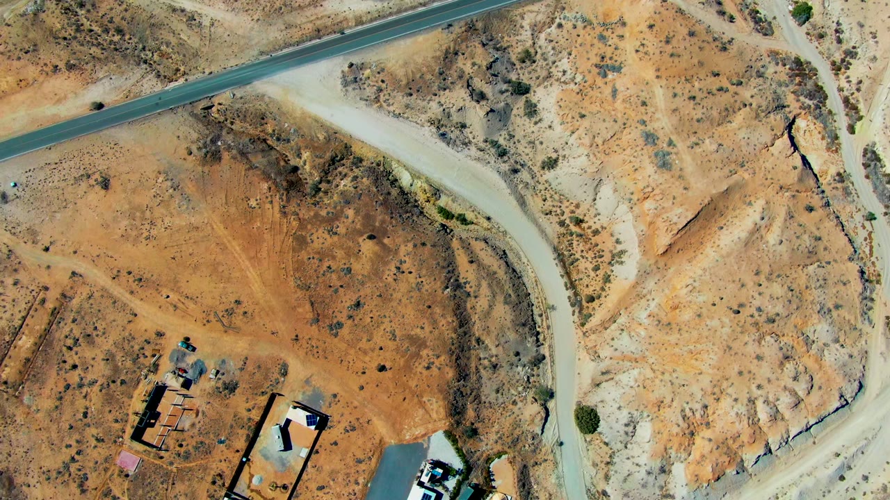 Aerial view over San Juanico desert terrain with pale dirt roads, a paved road at the top, scattered buildings near the bottom, small vehicles, and no visible people.