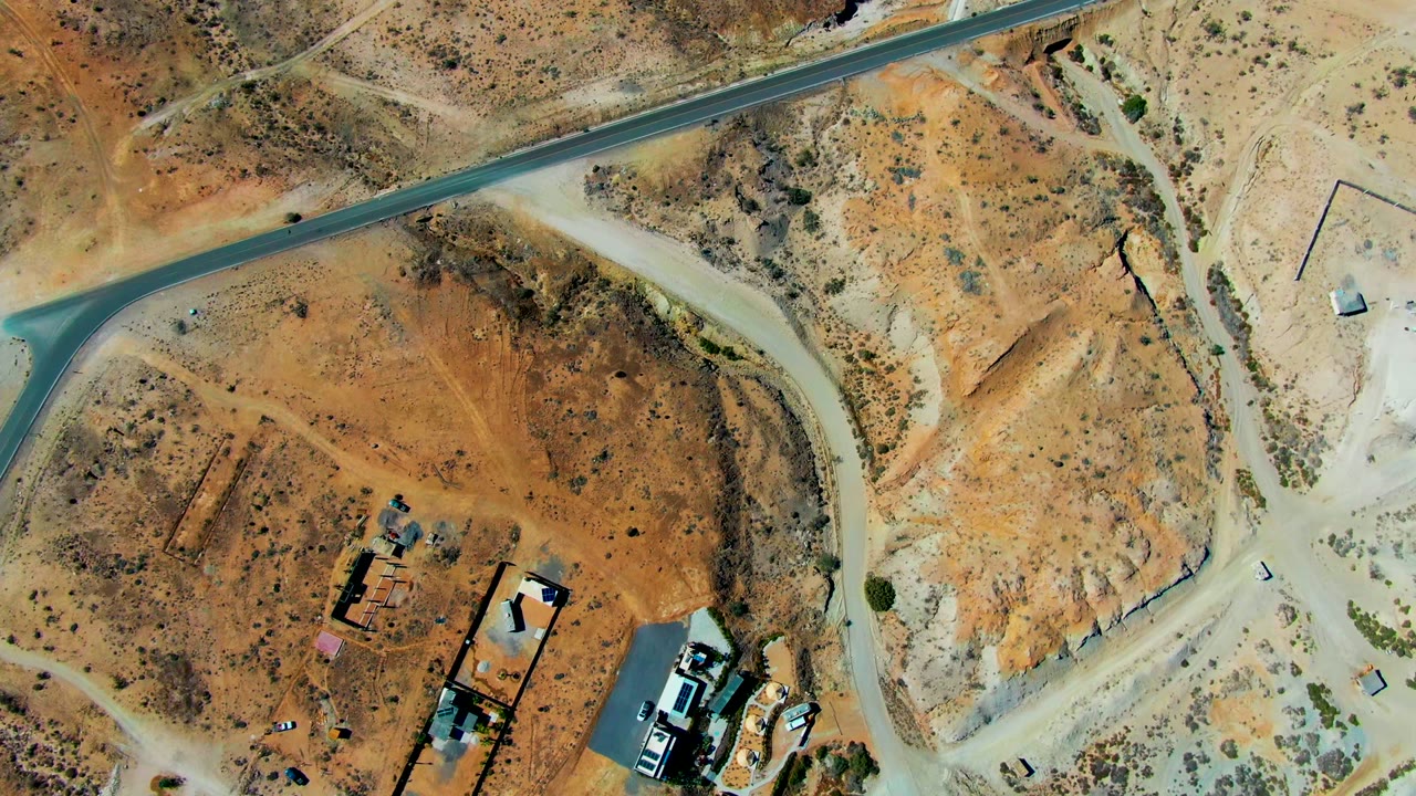 Top-down San Juanico frame with a pale dirt junction, rust-colored desert lots, several small vehicles near a building cluster, and no visible people.