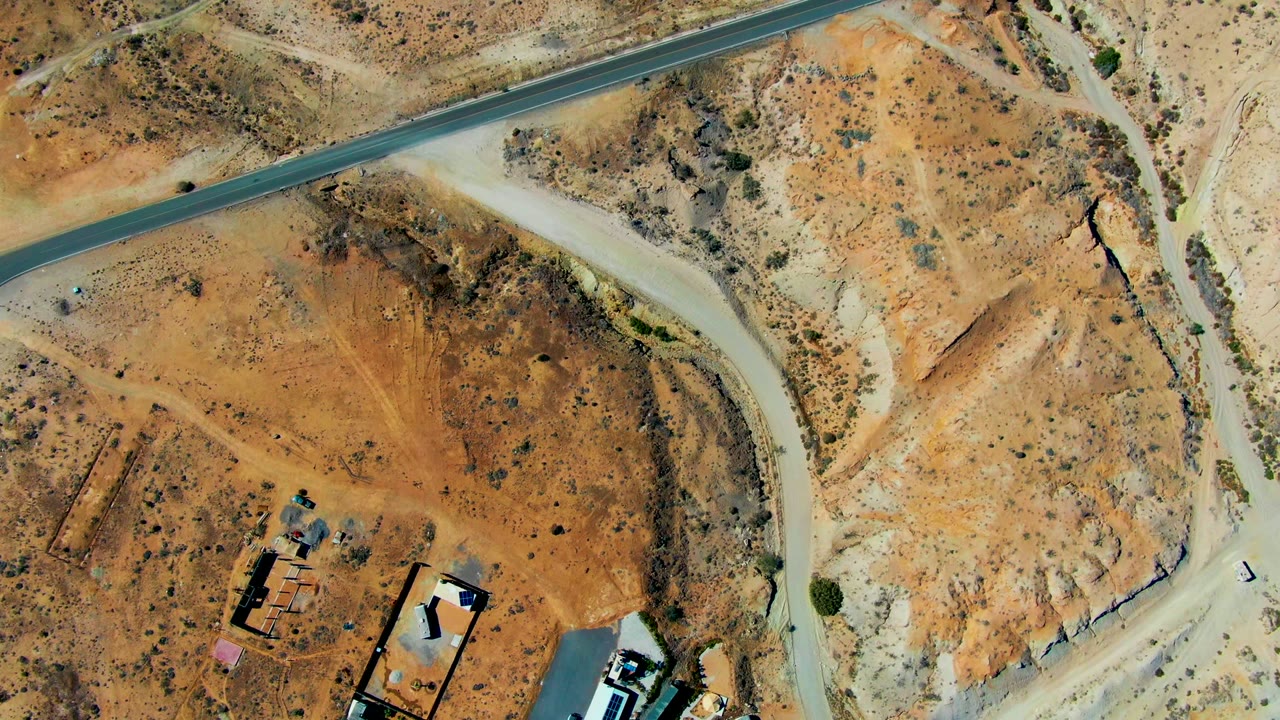 Overhead view in San Juanico, Baja California Sur showing a paved road at the top, a pale dirt road curving through brown desert terrain, small buildings at the lower left, and no visible people.