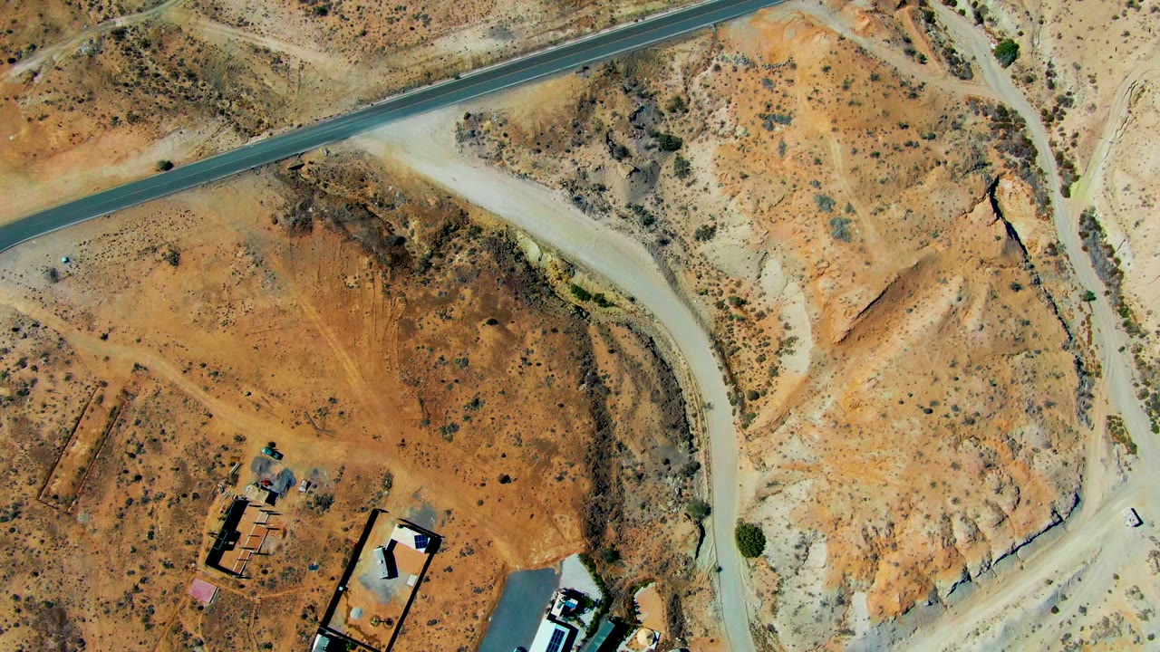 Aerial frame over San Juanico, Baja California Sur with the paved road running near the top, a curving dirt road crossing the center, scattered scrub, lower-edge buildings, and no visible people.