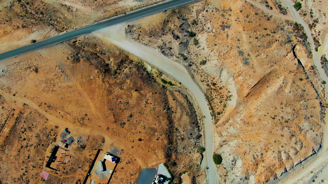 Top-down San Juanico desert view with a dark paved road along the upper frame, pale unpaved road bending below it, orange-brown terrain, sparse vegetation, and no visible people.