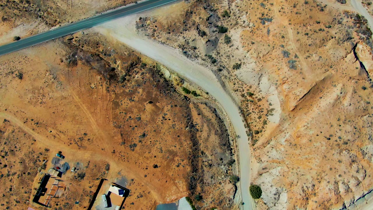 Overhead desert landscape with a curving dirt road and small buildings partly at the bottom edge.