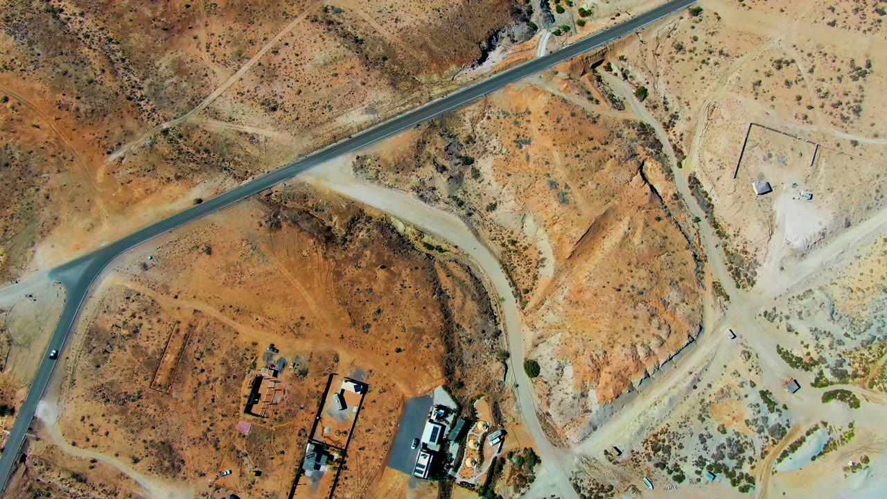 Aerial frame over San Juanico desert terrain showing the road bending past pale dirt tracks, rust-colored scrub, small buildings, vehicles, and no visible people.
