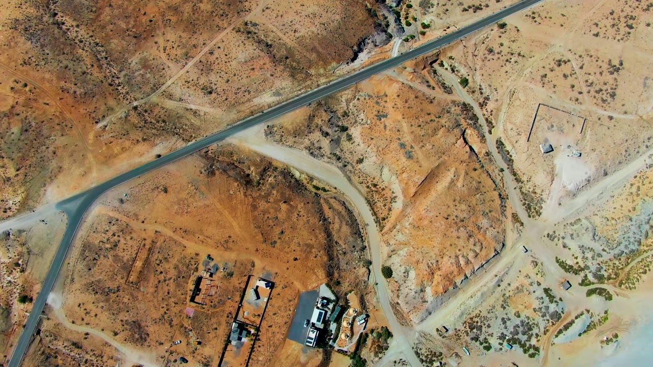 High aerial view over San Juanico desert terrain, showing a diagonal paved road, pale dirt tracks, small buildings, graded lots, and no visible people.