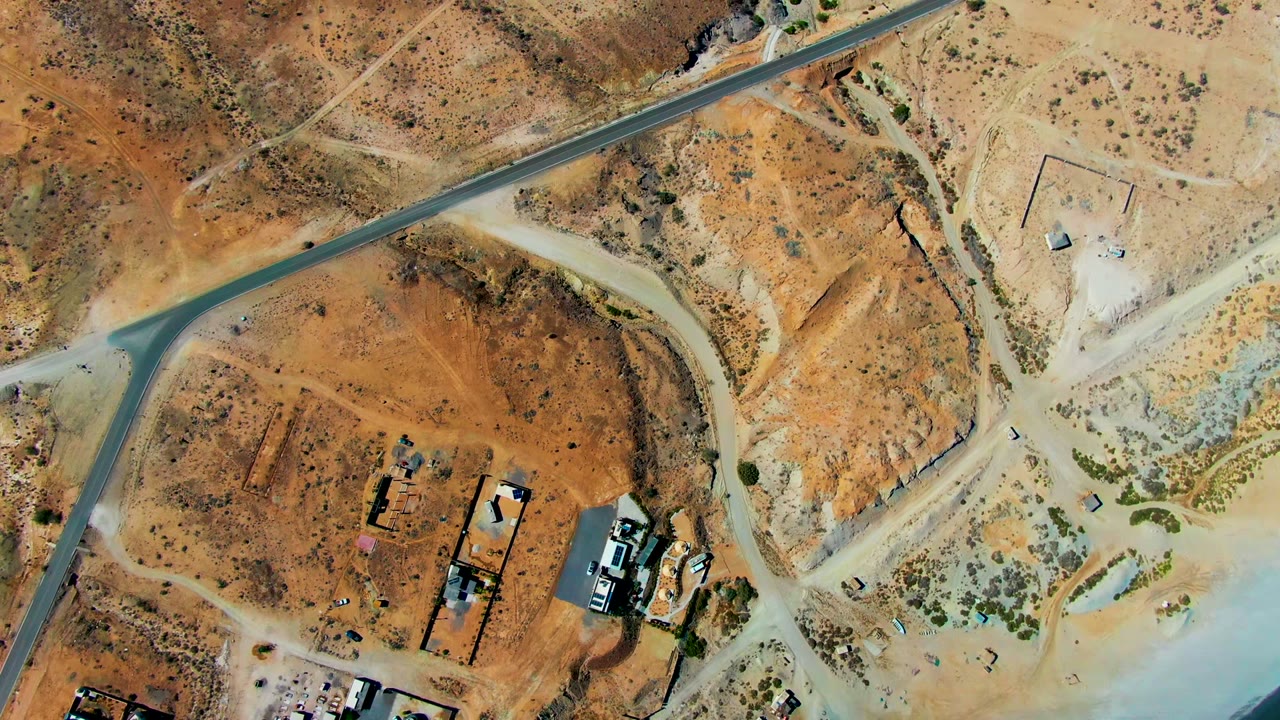Overhead view in San Juanico, Baja California Sur showing a diagonal paved strip, branching dirt roads, small construction structures, parked vehicles, tan desert ground, and no visible people.