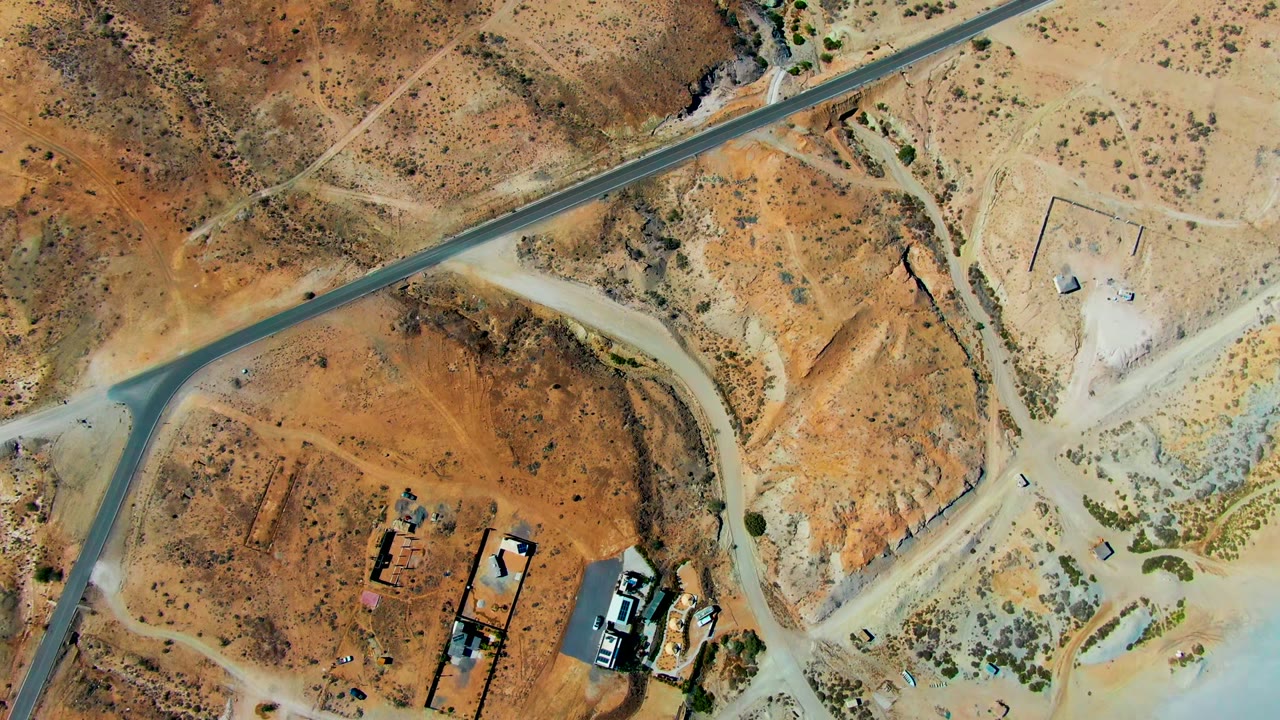 Top-down San Juanico, Baja California Sur view showing a diagonal runway or road, pale dirt tracks, a fenced or graded pad, scattered vehicles, brush patches, and no visible people.