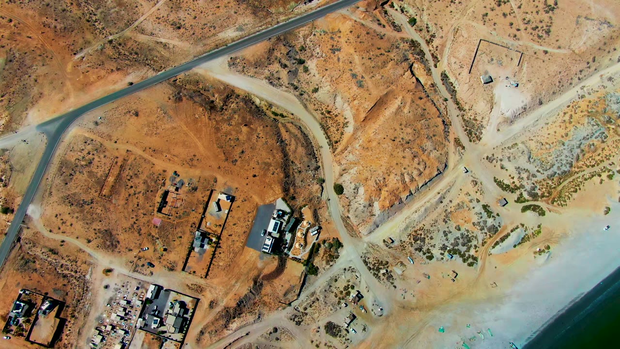 High overhead frame of San Juanico, Baja California Sur showing a paved road through sandy desert lots, scattered buildings, scrub, shoreline water, and no visible people.