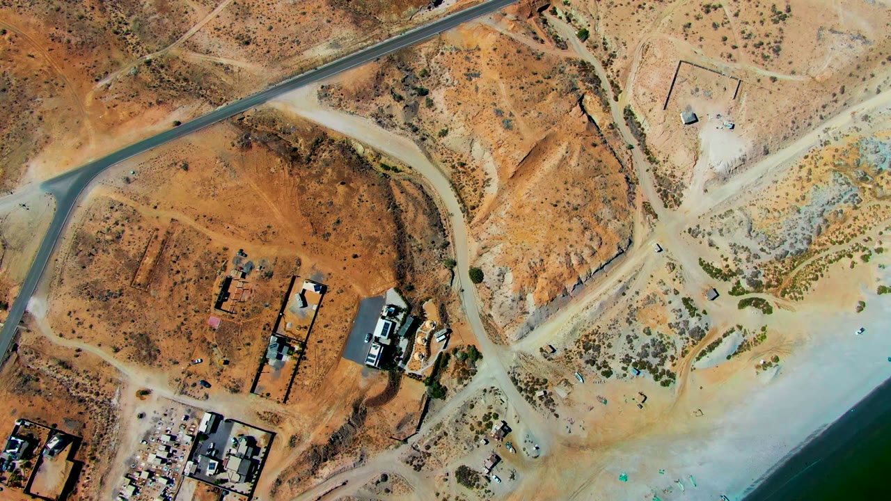 Aerial view of a paved road crossing arid terrain near small structures and water.