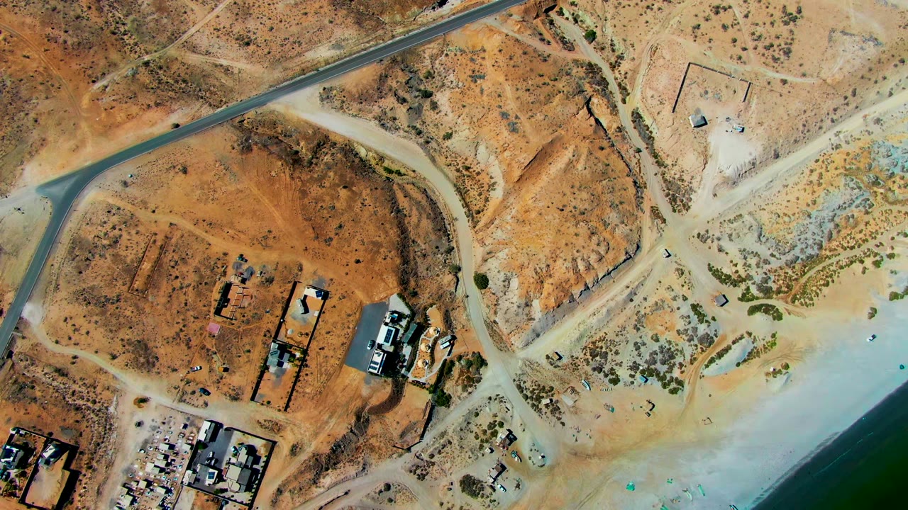 Top-down frame over San Juanico with the road near the upper edge, open sandy lots, low buildings, pale shoreline water, and no visible people.