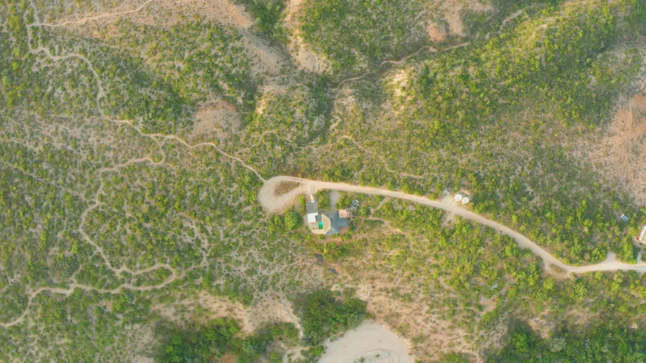 Overhead frame from Cabo Pulmo, Baja California Sur showing a pale dirt road, green-brown hillside vegetation, small roadside structures, and no visible people.